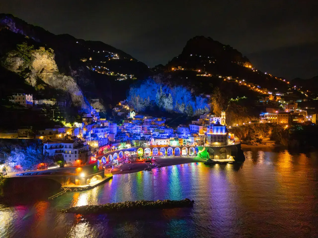 A vibrant night view of Vietri sul Mare, with buildings illuminated in colorful lights reflecting on the water.