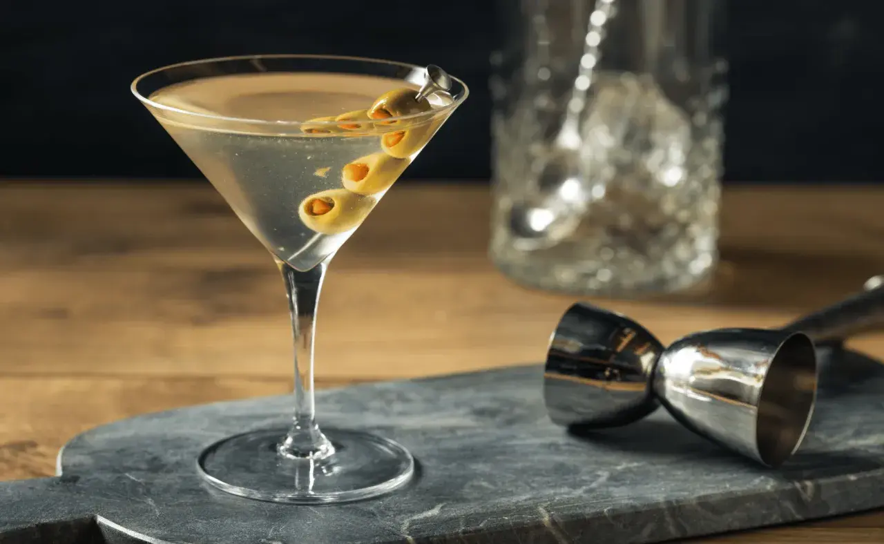 Un verre de martini gin, garni de trois olives, prêt à être dégusté. Un doseur à cocktail et un pichet à glaçons sont visibles en arrière-plan.