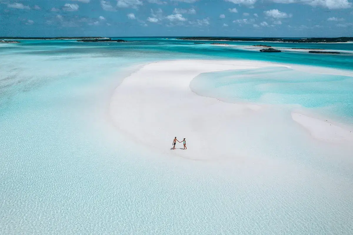 Un couple se tient la main sur une plage de sable blanc aux Bahamas. L'eau turquoise et les îlots au loin invitent à explorer que faire aux Bahamas.