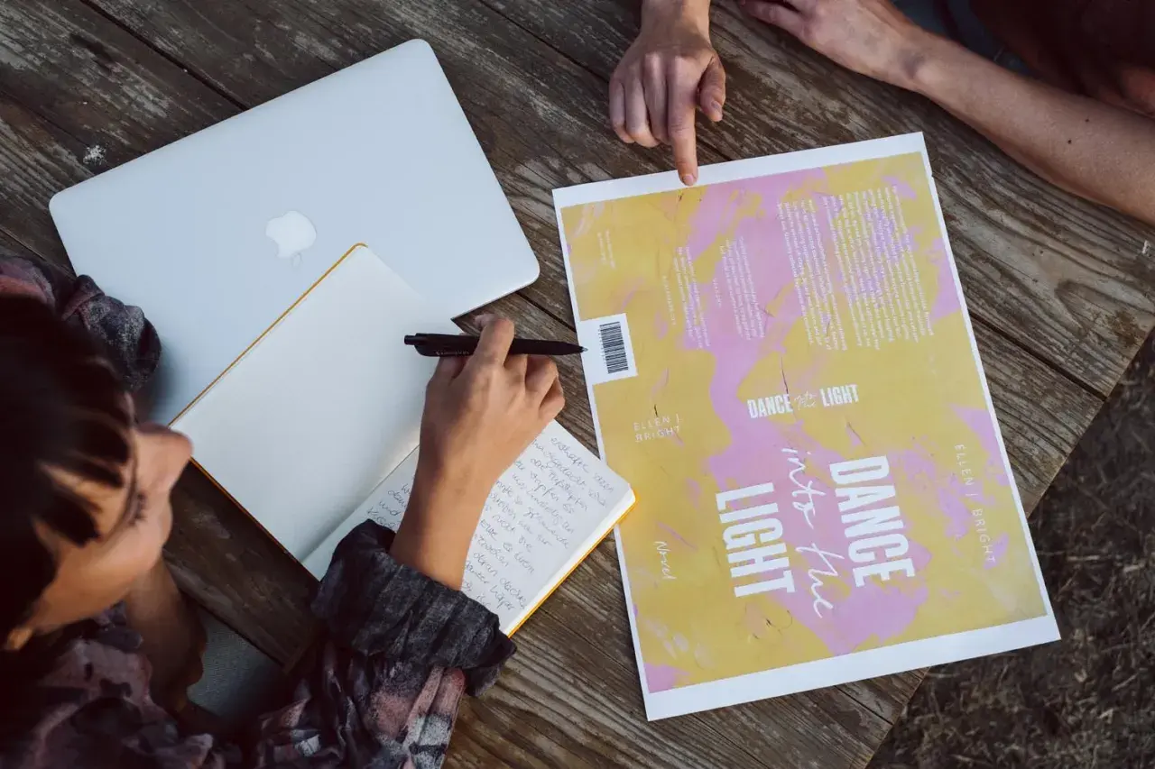 Des mains travaillent à créer une couverture de livre. Un ordinateur portable, un carnet et un brouillon de couverture sont sur une table en bois.