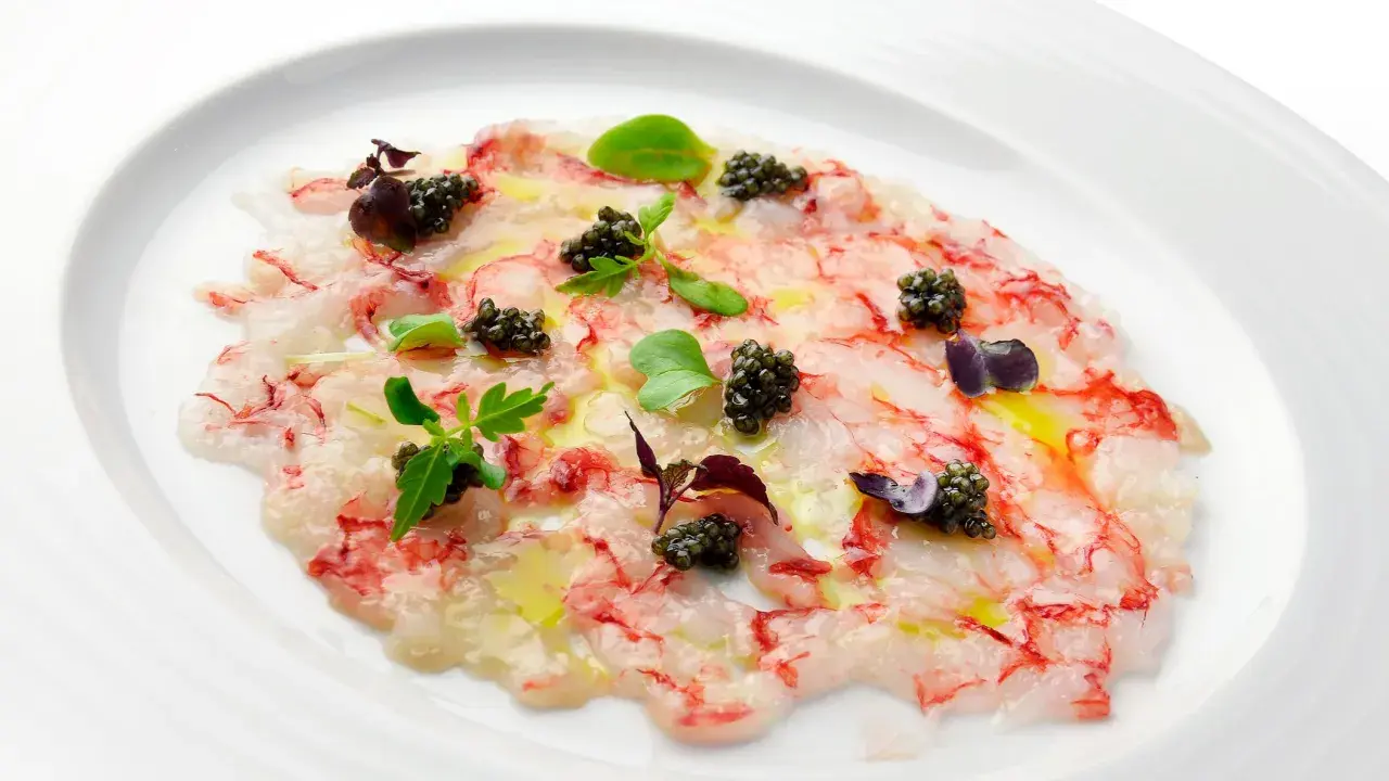 Delicado carpaccio de gambas con caviar, aceite de oliva y brotes verdes, presentado en un plato blanco.