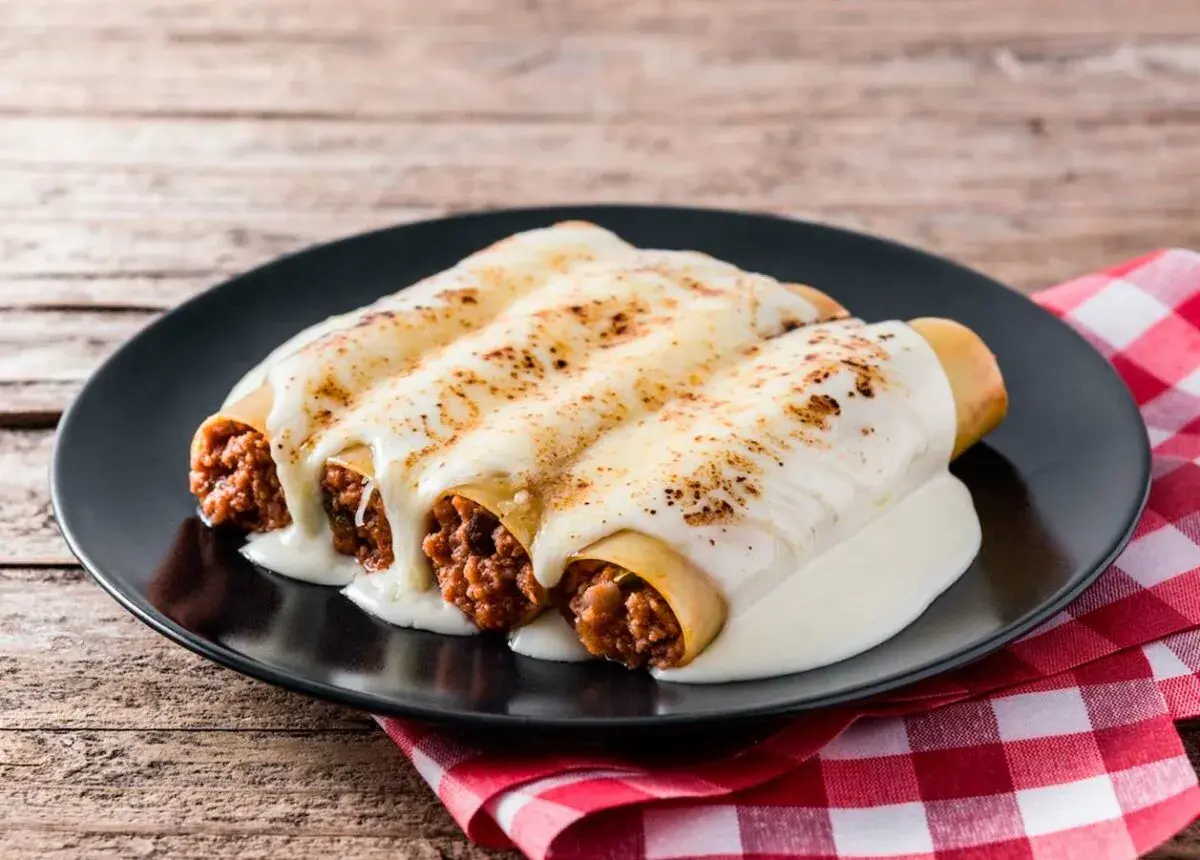 Tres canelones de carne gratinados con queso cremoso, servidos en un plato negro sobre un mantel a cuadros rojos.