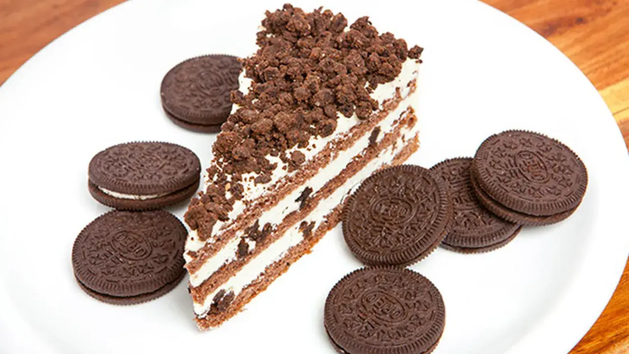 Rebanada de bizcocho de oreo con capas de crema y galleta triturada, rodeada de galletas oreo enteras en un plato blanco.