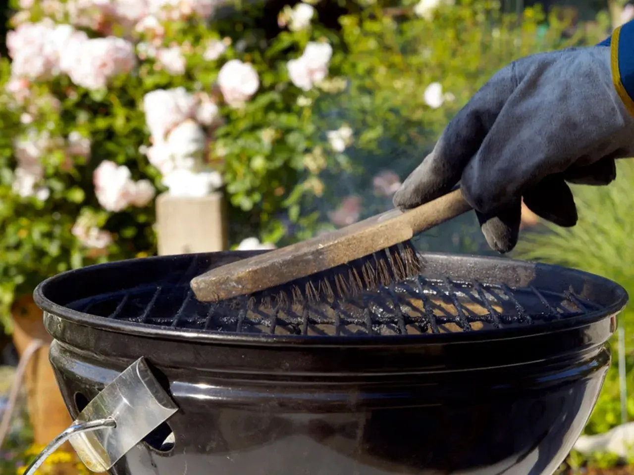 Main tenant gantée, brosse métallique pour nettoyer un barbecue. Des roses roses en arrière-plan.