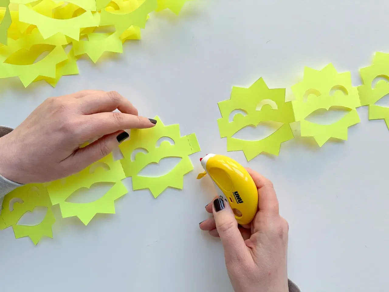 Hände basteln eine ziehharmonika-girlande aus gelben Sonnen mit Gesichtern. Ein Klebestift verbindet die Elemente.