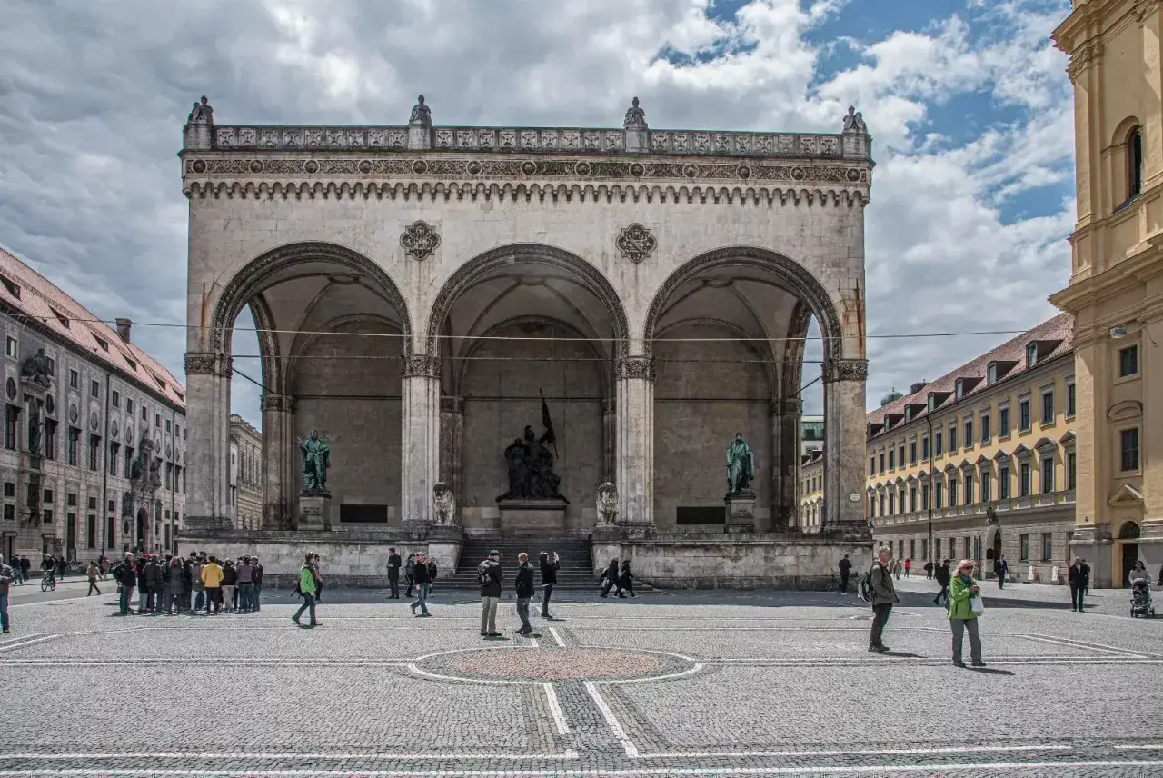 Der Odeonsplatz in München mit der Feldherrnhalle und ihren Arkaden. Menschen schlendern über den Platz, während die Sonne durch die Wolken bricht.