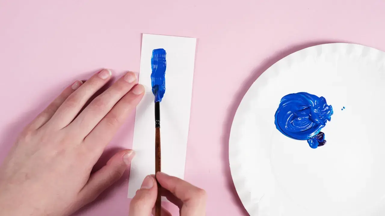Eine Hand hält einen Pinsel, um mit leuchtendem Blau auf weißem Papier zu malen. Daneben auf einer Pappplatte wird dunkelblau gemischt.