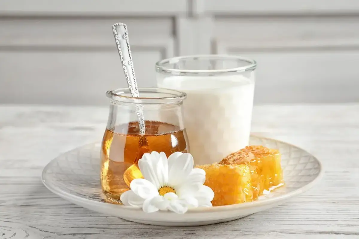 Un bicchiere di latte, un vasetto di miele dorato con un cucchiaino, e favi di miele su un piatto con un fiore bianco. Alcuni credono che latte caldo e miele faccia male.