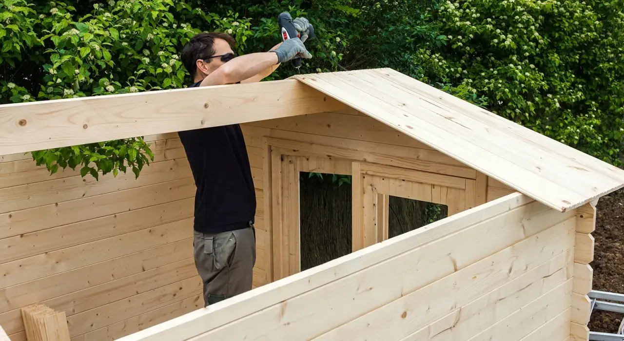 Un homme assemble une cabane en bois, utilisant une perceuse pour fixer une planche sur le toit. La peinture sur bois est visible sur les murs et le toit.