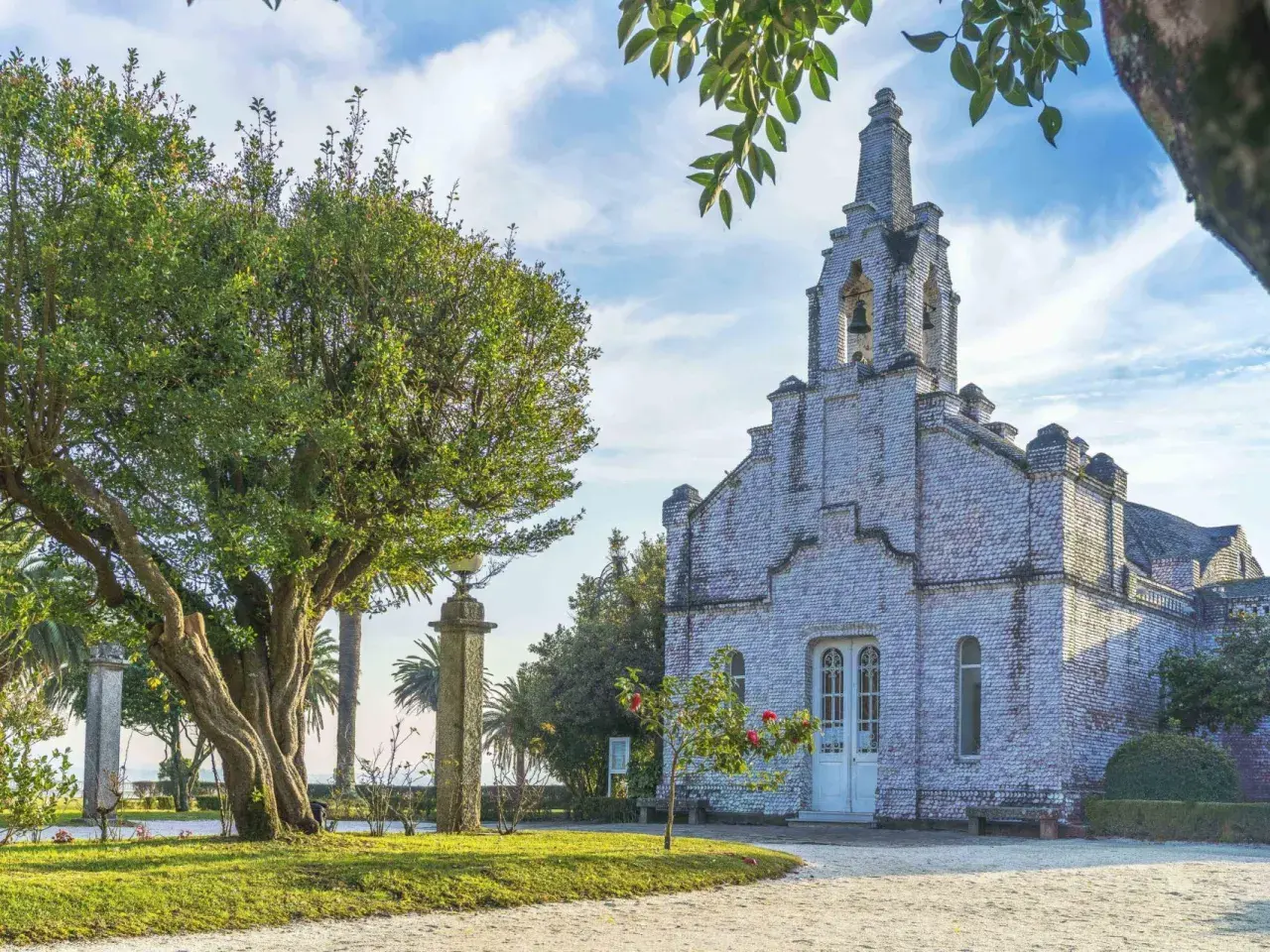 La iglesia de la Toja, cubierta de conchas, se alza majestuosa bajo un cielo azul con nubes. Un árbol frondoso y palmeras la rodean.