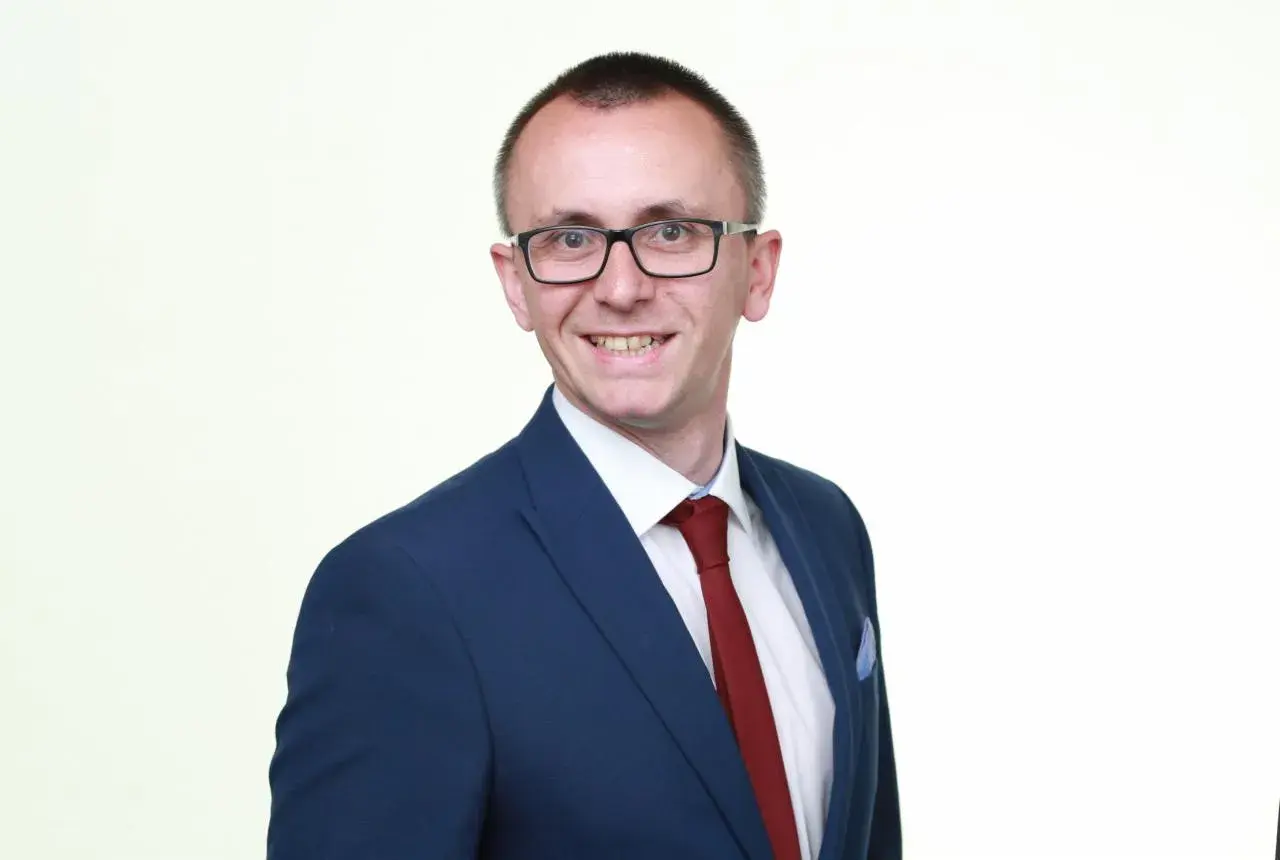 Mężczyzna w okularach i granatowej marynarce z czerwonym krawatem, uśmiecha się.