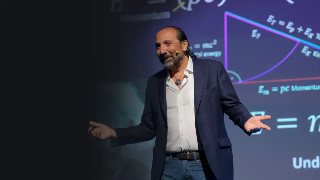 Nassim Haramein, con gestos amplios, explica conceptos de física ante una pantalla con fórmulas como E=mc².