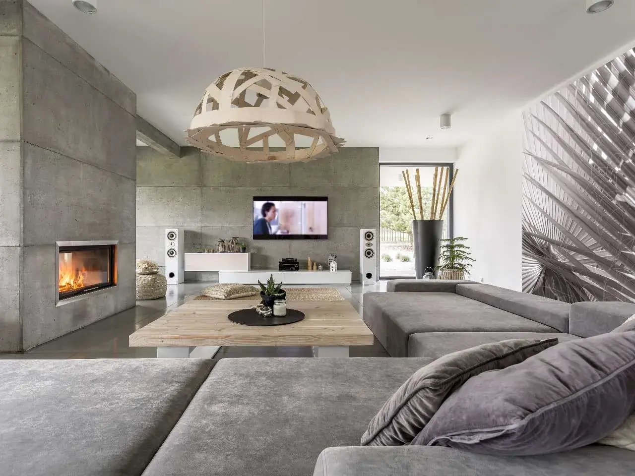 Nowoczesny salon z kominkiem i ścianą z betonu architektonicznego. Szara kanapa, drewniany stolik i designerska lampa tworzą przytulną atmosferę.