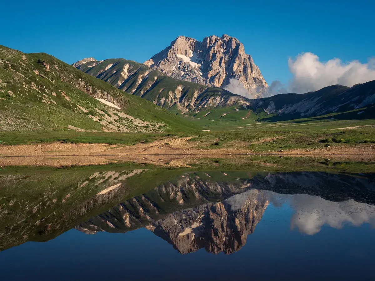 Spiegelglatte See reflektiert majestätisches **Gebirge in Italien** unter blauem Himmel. Grüne Hänge und felsige Gipfel.