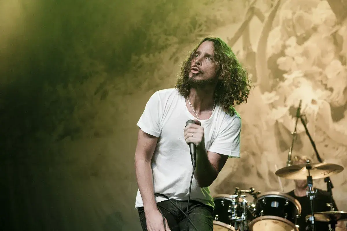 Chris Cornell z zespołem na scenie, w białej koszulce, śpiewa do mikrofonu. W tle perkusja i światła.