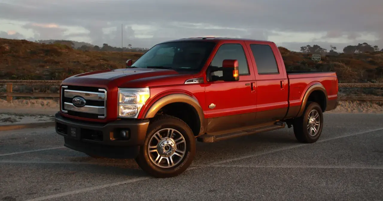 Czerwony Ford F350 King Ranch z brązowym pasem, zaparkowany na tle krajobrazu z wydmami i drewnianym płotem.