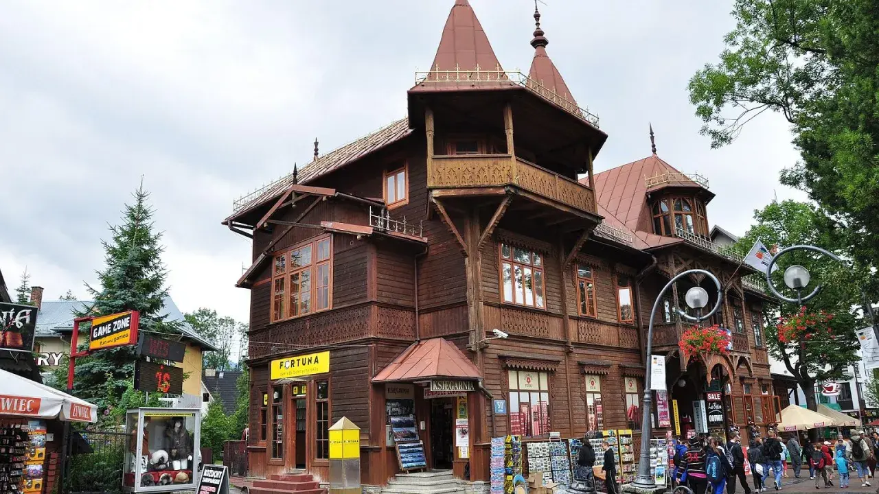 Drewniana architektura na Krupówkach, z charakterystycznymi zdobieniami i wieżyczkami. W tle widać ludzi spacerujących po tętniącej życiem ulicy.