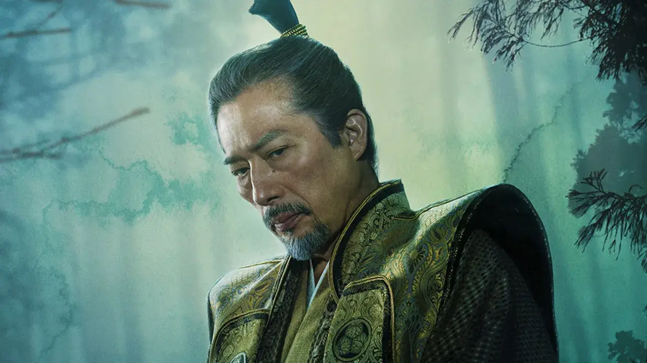 En äldre man med gråsprängt skägg och en tofs i håret, klädd i en utsmyckad dräkt. En karaktär från shogun serie.
