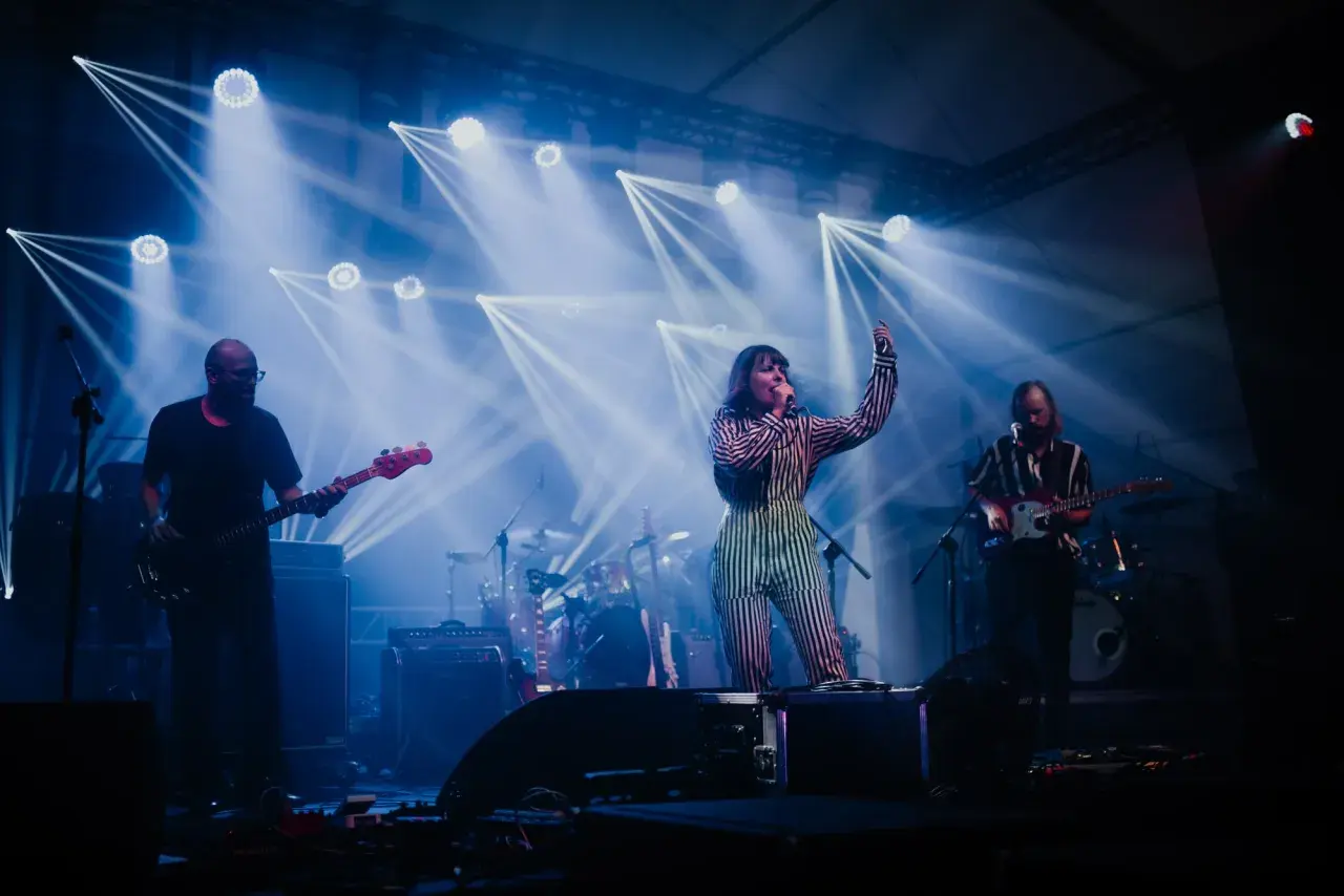 Izzy i The Black Trees na scenie, otoczeni niebieskimi światłami. Wokalistka w pasiastym stroju śpiewa, gitarzyści grają.