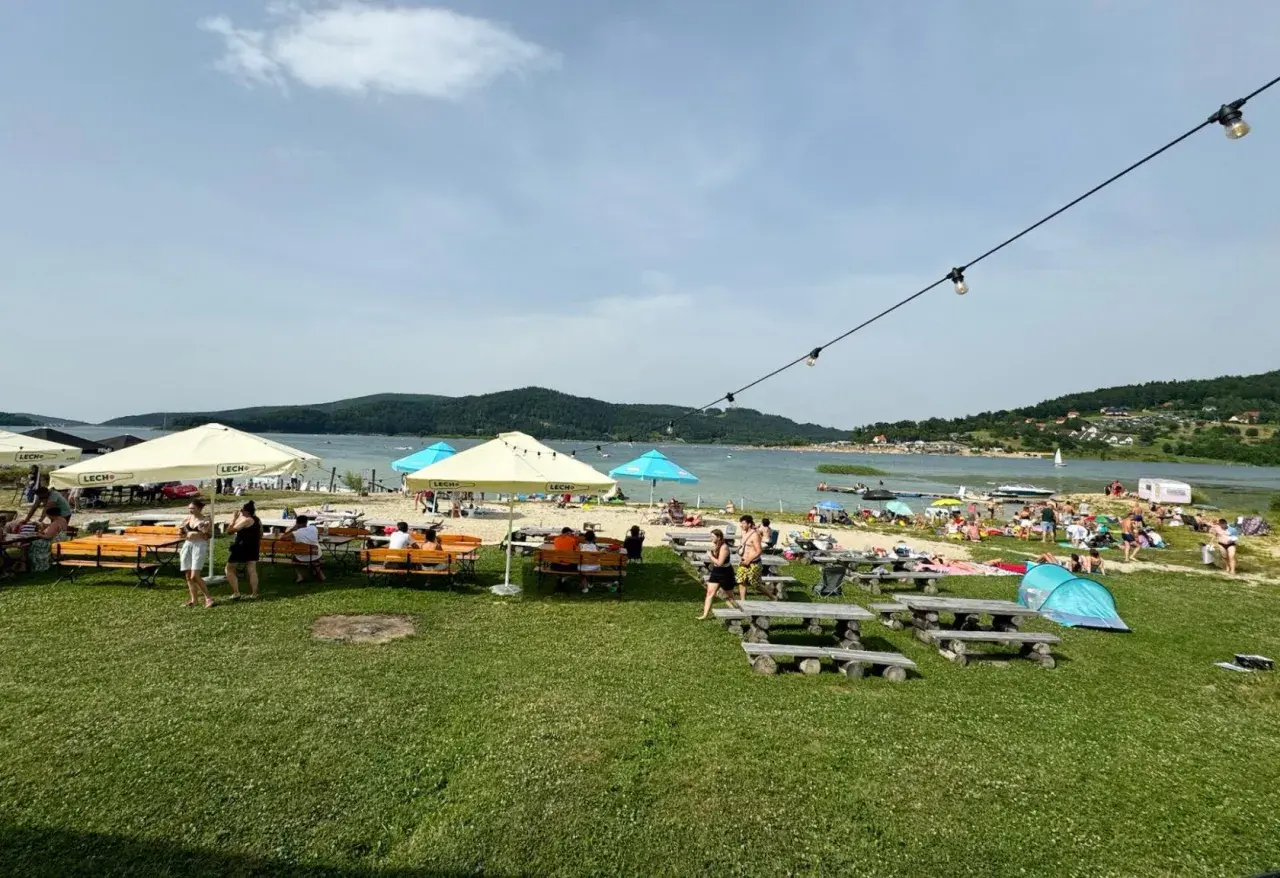 Słoneczny dzień nad Jeziorem Mucharskim. Ludzie odpoczywają na plaży, pod parasolami, przy stolikach. W tle jezioro i wzgórza. Atrakcje dla każdego.