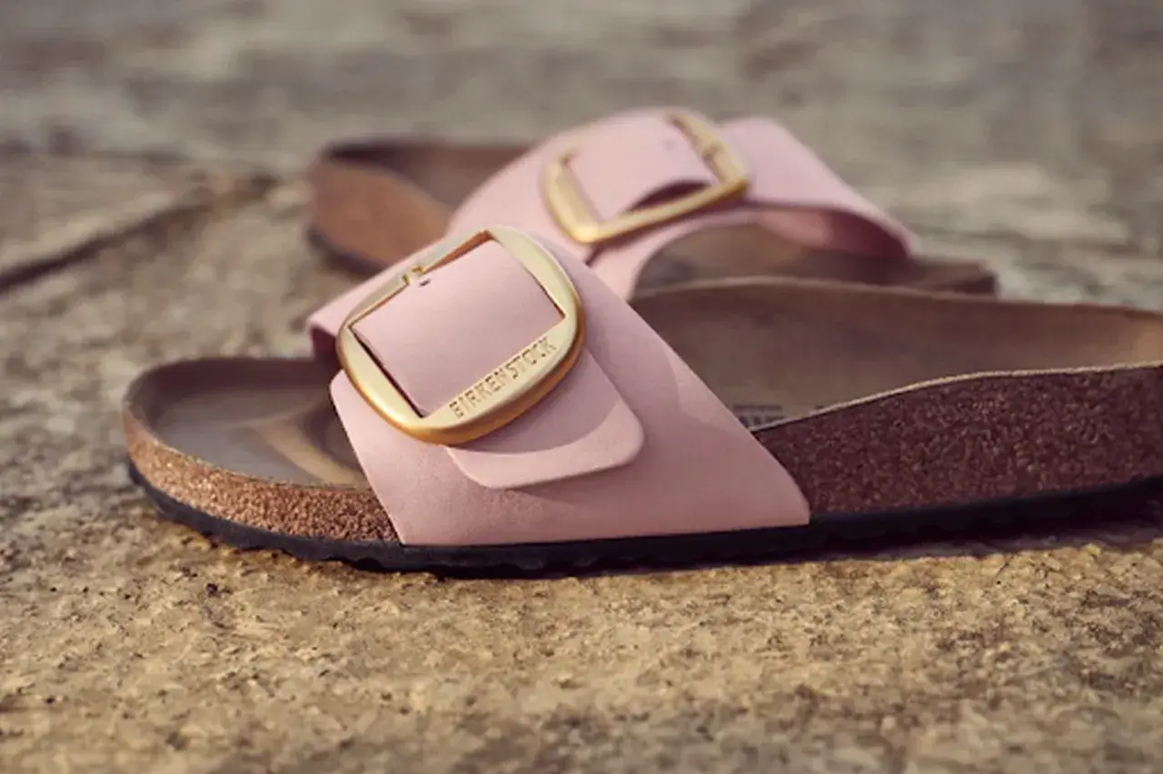 Różowe klapki Birkenstock z złotą klamrą, leżące na kamiennym podłożu.