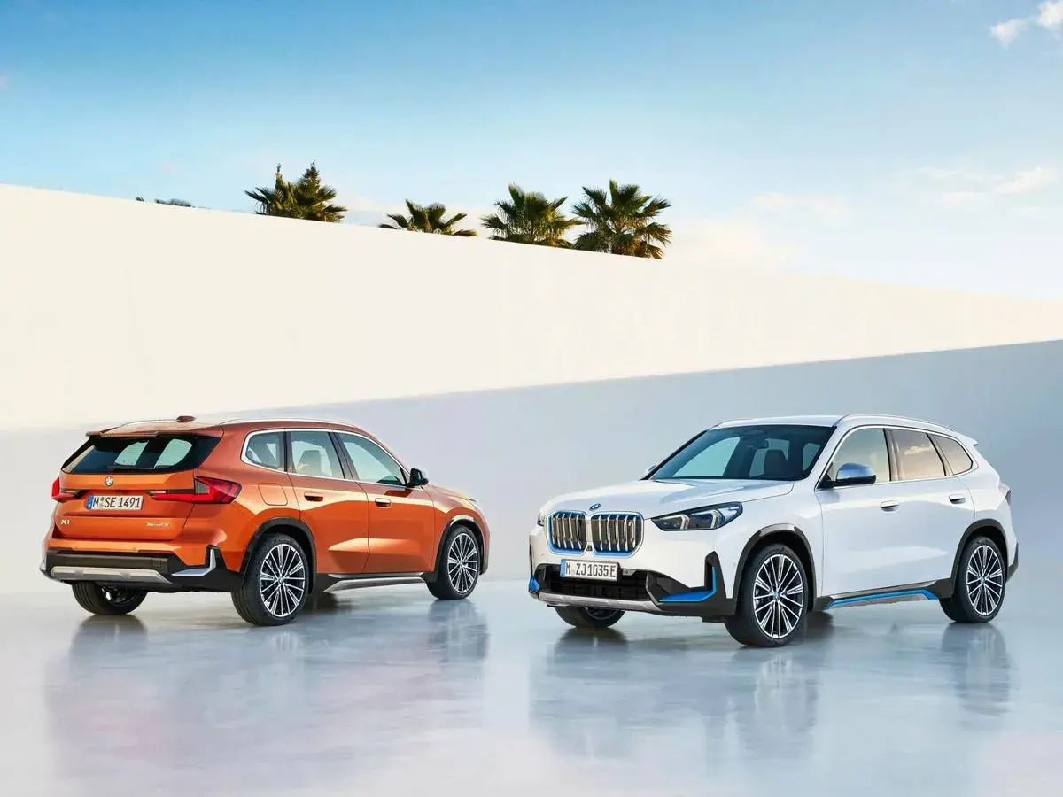 Dos BMW Serie X, uno naranja y otro blanco, posan ante un muro blanco y palmeras.