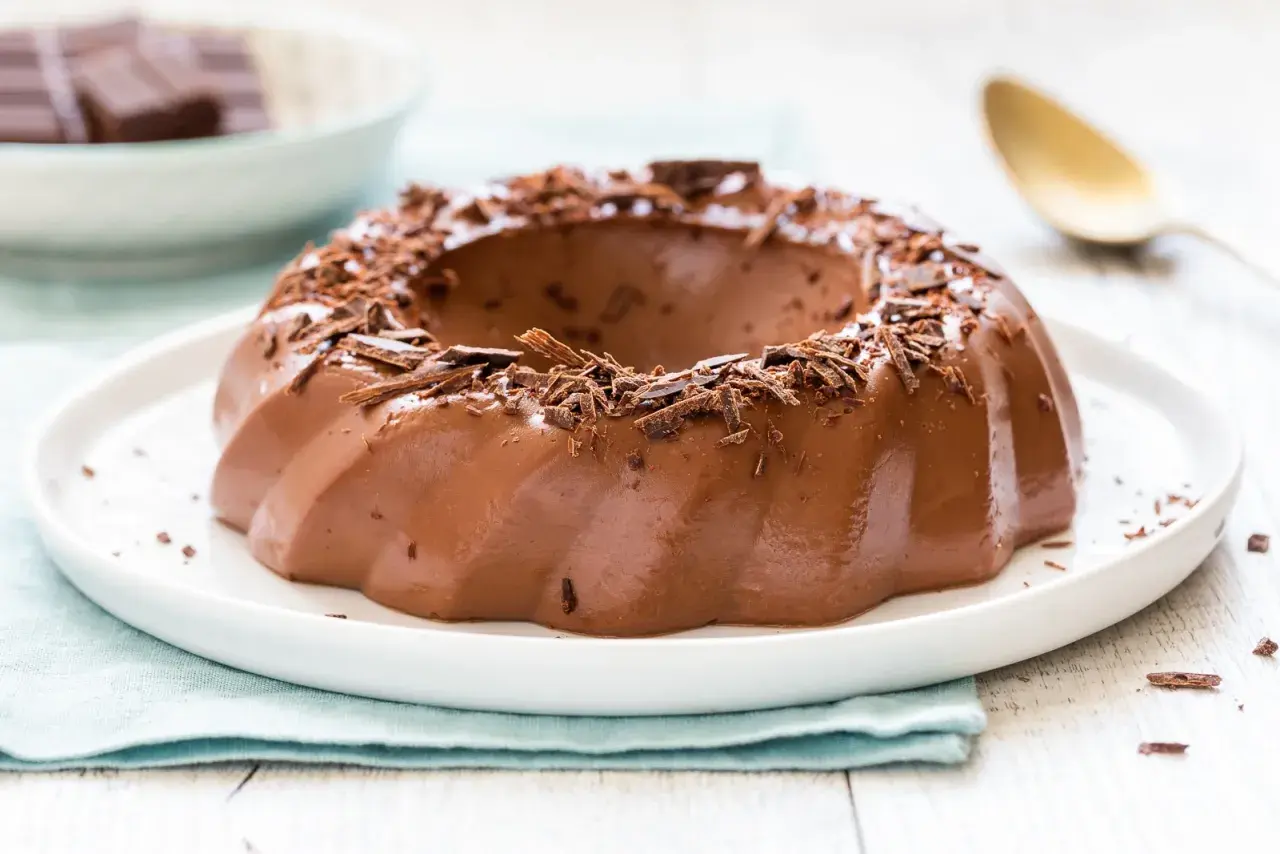 Budino al cioccolato lucido e invitante, guarnito con riccioli di cioccolato fondente, su un piatto bianco.