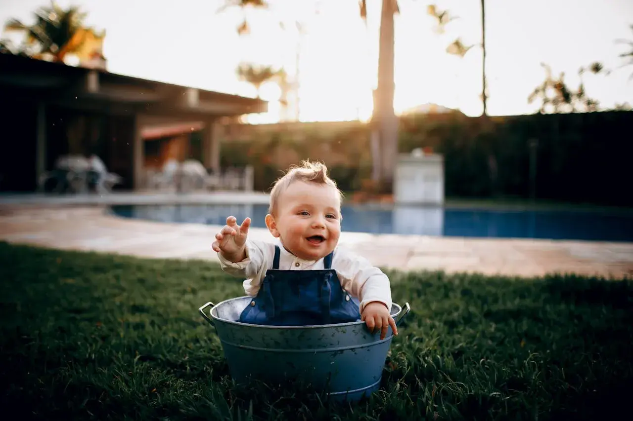 Glad bebis i en hink på gräsmattan, med en pool i bakgrunden. En härlig dag för ett bebis bada i poolen!