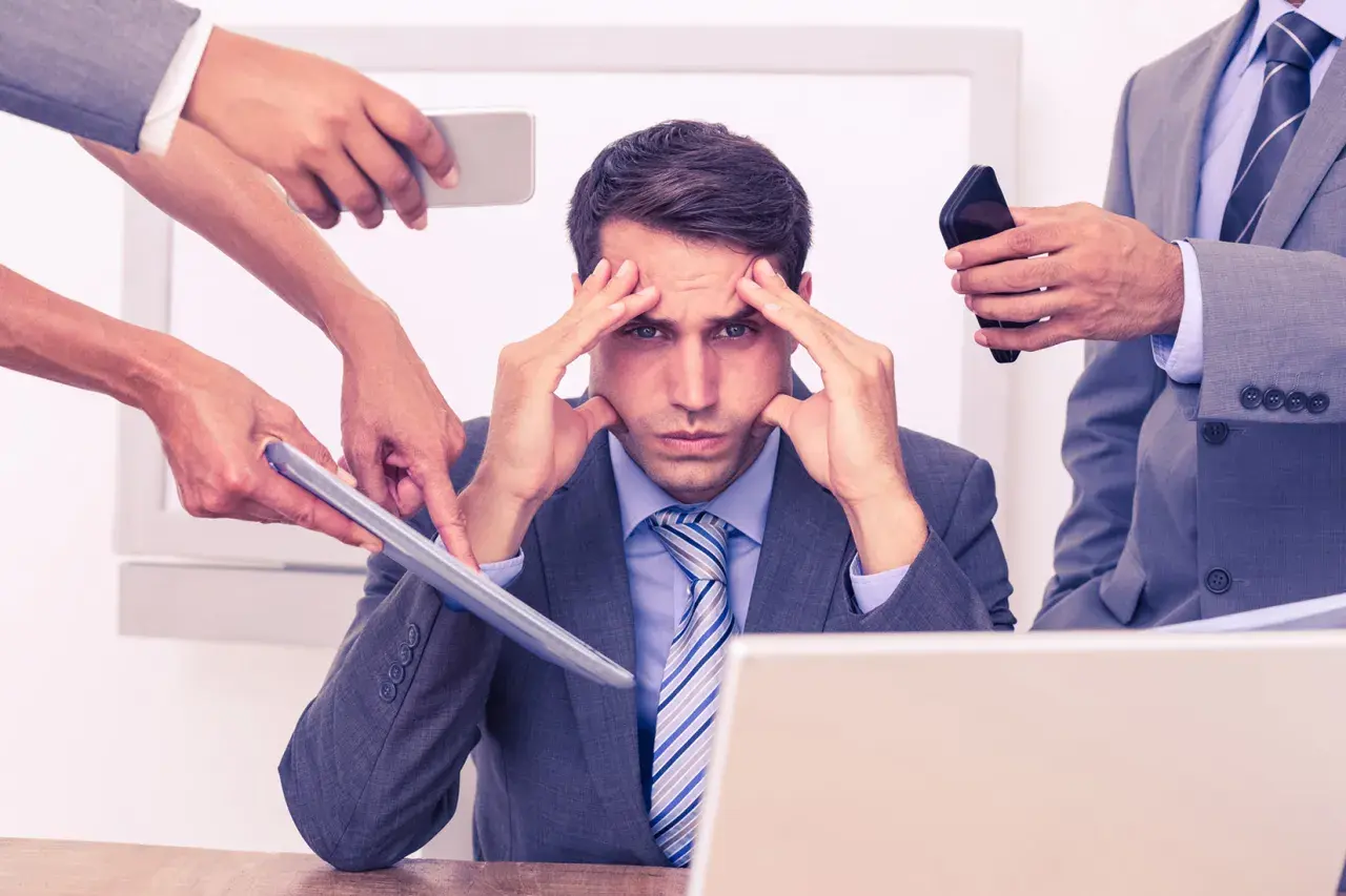 Stressad man med händer på tinningarna, omgiven av kollegor som ger instruktioner. Dålig kognitiv ergonomi leder till överbelastning.