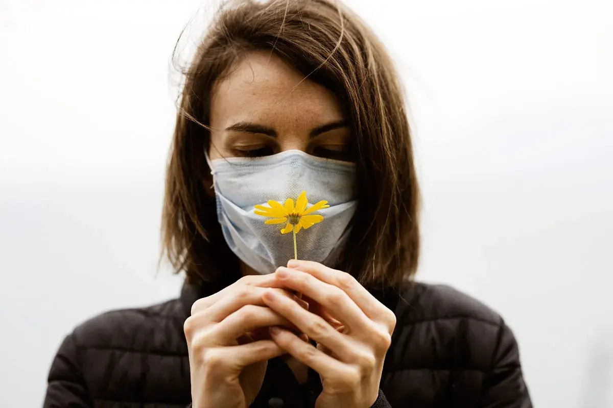 Mujer con mascarilla azul, oliendo una flor amarilla. Diversos tipos de mascarillas protegen, pero la esperanza florece.