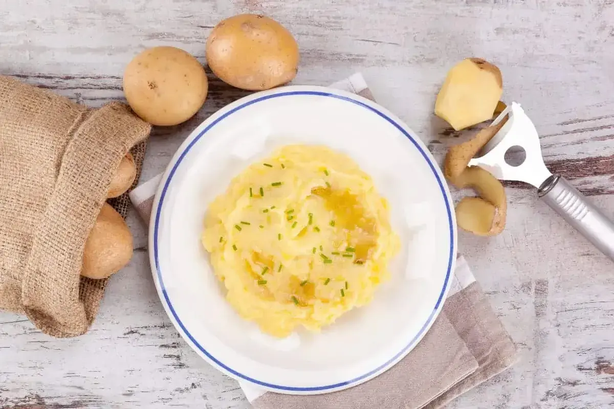 Pyszne puree ziemniaczane na talerzu, obok surowe ziemniaki i obieraczka. Czy cukrzyk może jeść ziemniaki? Tak, w umiarkowanych ilościach!
