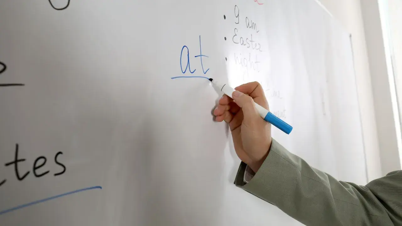 Mano scrive "at" su lavagna, spiegando il suo significato in inglese.