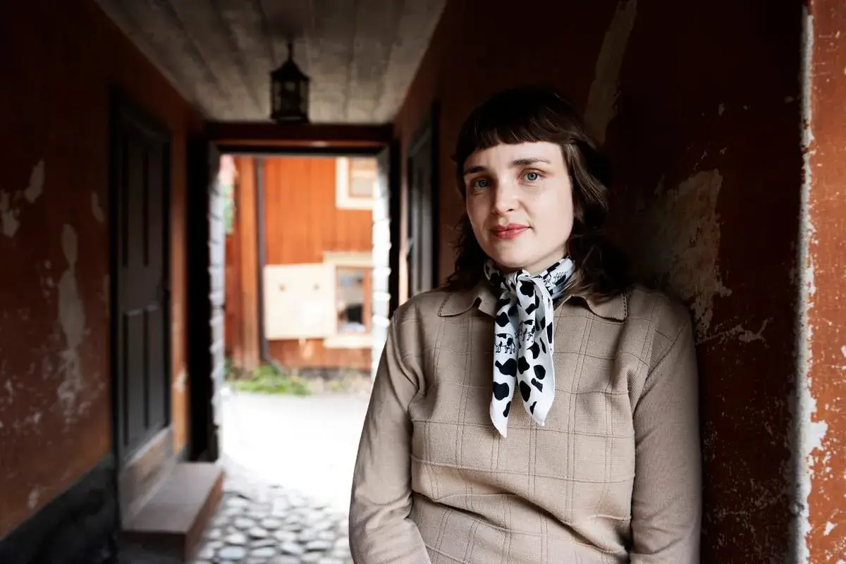 Sanna Samuelsson står i en gång med orange väggar och en träfasad i bakgrunden. Hon bär en beige tröja och en scarf med koskinnmönster.