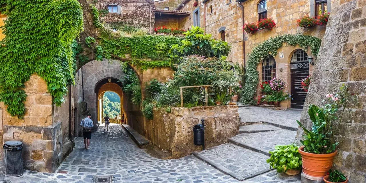 Un vicolo acciottolato in un borgo da visitare, con case in pietra ricoperte di edera e fiori. Un arco conduce a una vista panoramica.