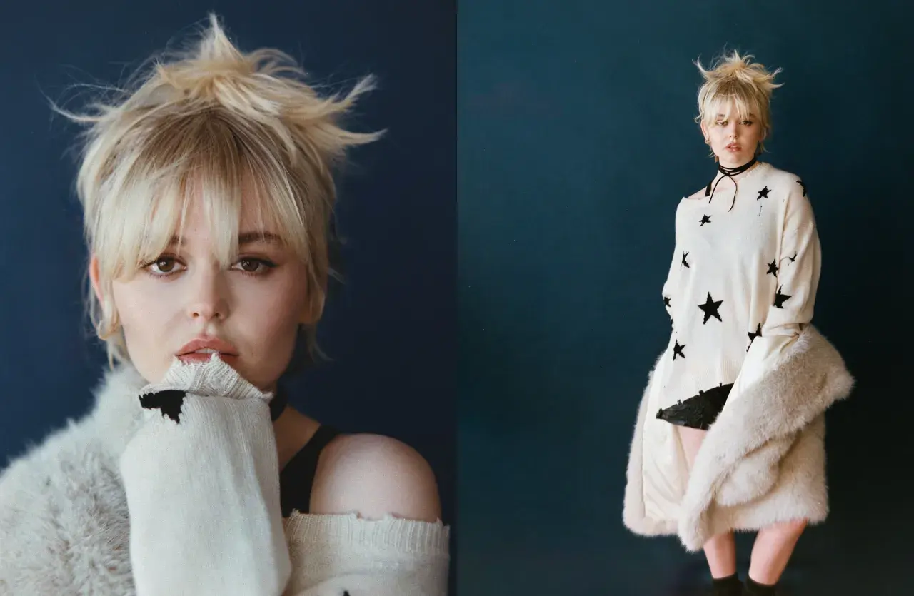 Emily Alyn Lind med rufsigt blont hår, bär en stickad tröja med stjärnor och en pälskappa.