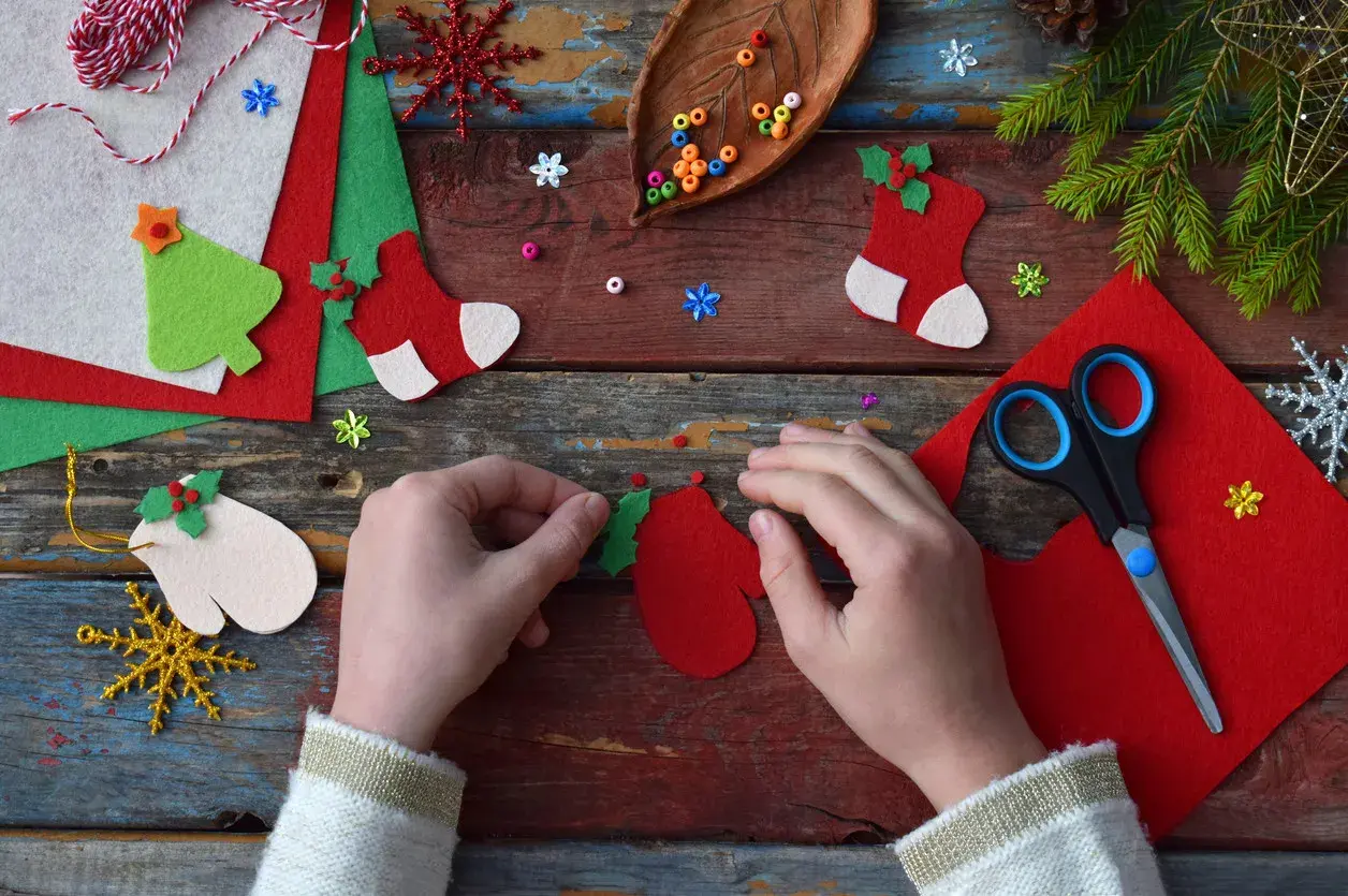 Manos creando adornos navideños con fieltro rojo y verde. Tijeras, cuentas y piñas completan la escena de manualidades.