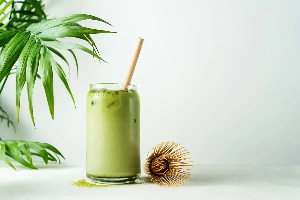 Orzeźwiające matcha latte z lodem i bambusową słomką, obok bambusowa miotełka. Poznaj niezwykłe matcha właściwości!