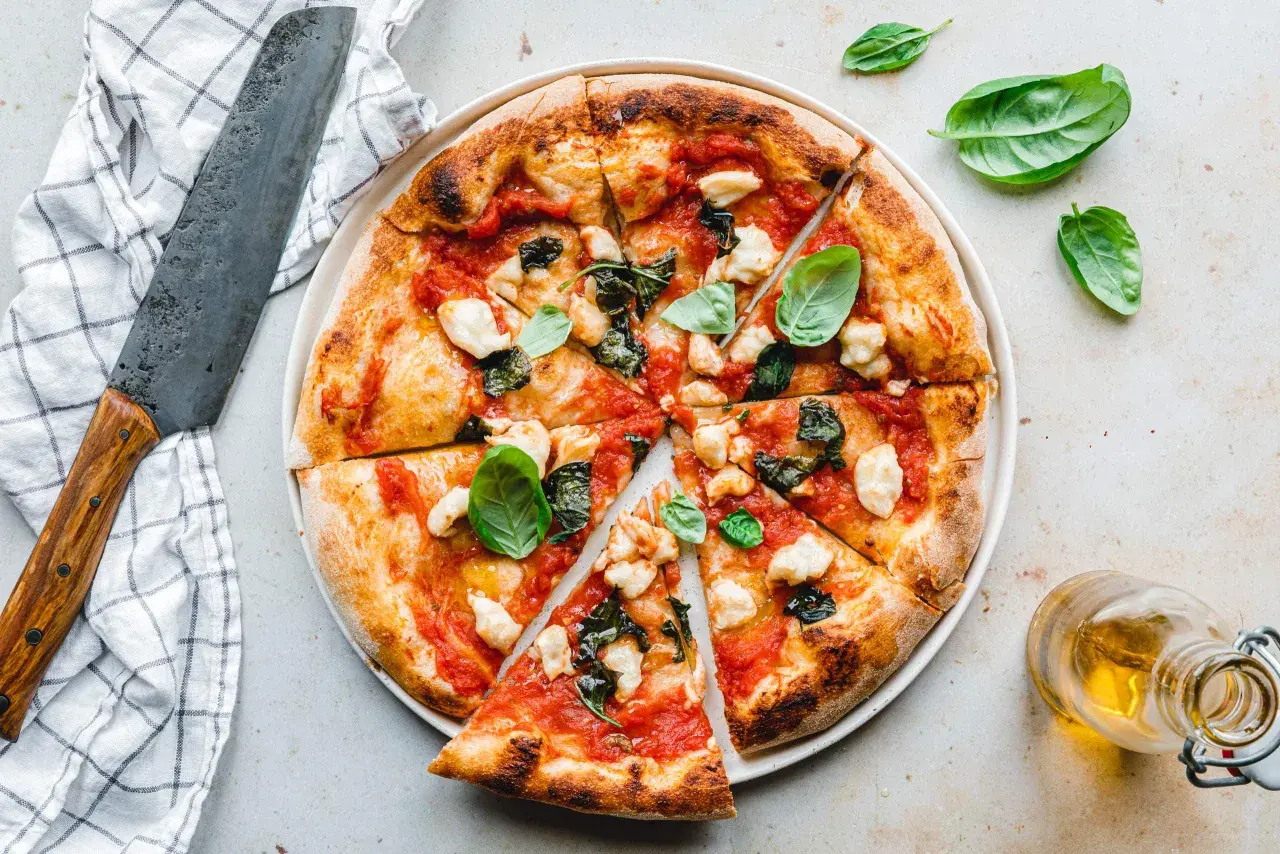 Eine perfekt gebackene vegane Pizza mit Tomaten, veganem Mozzarella und Basilikum, daneben ein Messer und Olivenöl.