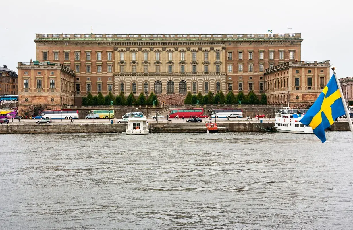 Stor byggnad vid vattnet, troligen kuninkaanlinna tukholma, med bussar och båtar. Svenska flaggan vajar.