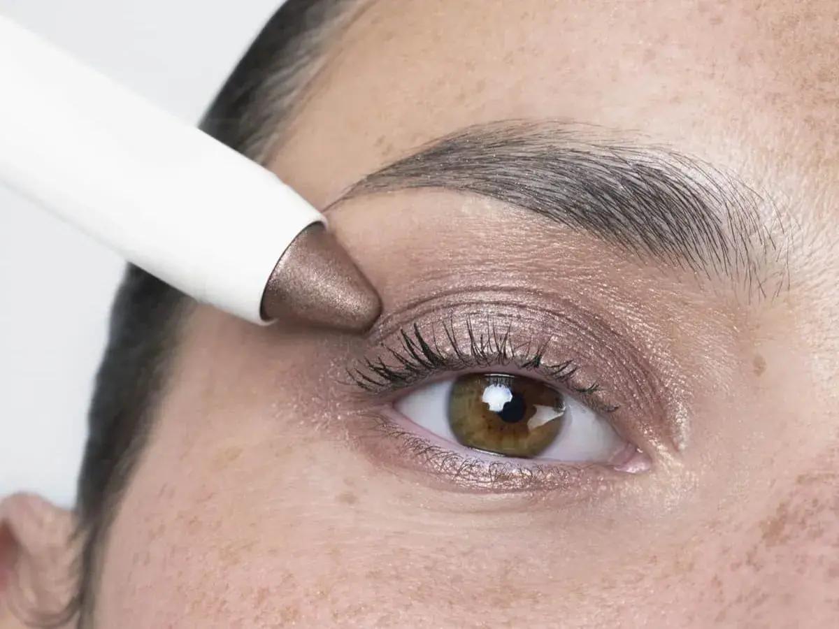 Application d'un fard à paupières bronze sur des yeux marrons. Un bâtonnet blanc dépose la couleur sur la paupière.