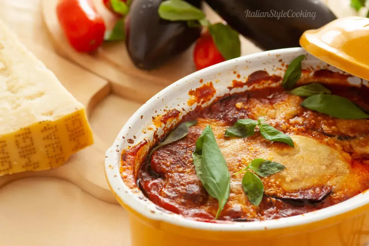 Gebackene Auberginen mit Tomatensoße und Basilikum in einer Auflaufform, daneben Parmesan und frische Auberginen.