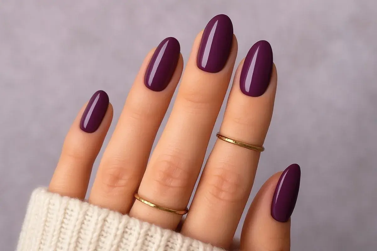 Main d'une femme aux ongles vernis d'un prune profond, une parfaite idée ongle janvier. Deux fines bagues dorées ornent les doigts.