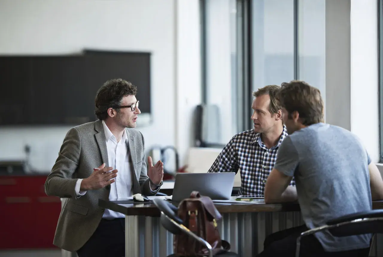 A man in a blazer gestures animatedly, a positive change agent discussing ideas with colleagues at a modern office table.
