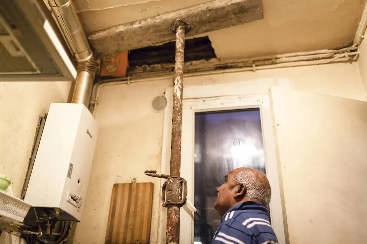 Un homme regarde un plafond délabré soutenu par une poutre rouillée. Ce **logement insalubre** montre des signes de dégradation avancée.