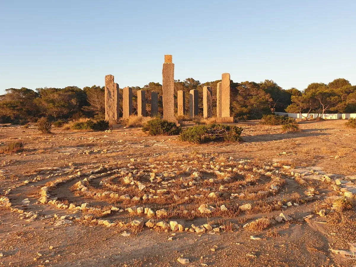 Steine bilden ein Labyrinth im Freien, umgeben von Pinien. Im Hintergrund ragen steinerne Säulen auf, die an Zeit und Raum auf Ibiza erinnern.