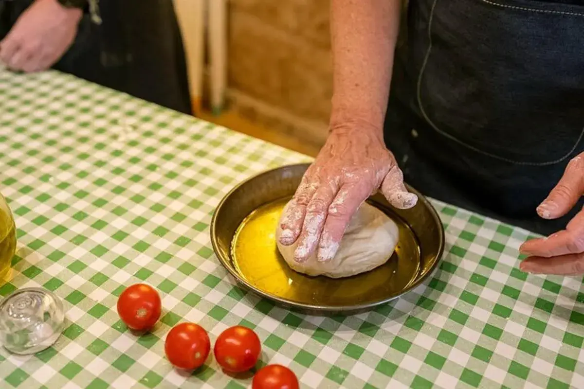 Ręce w mące wyrabiają ciasto na oliwie. Na zielono-białej kratce leżą pomidorki. Pyszne bari jedzenie w przygotowaniu.