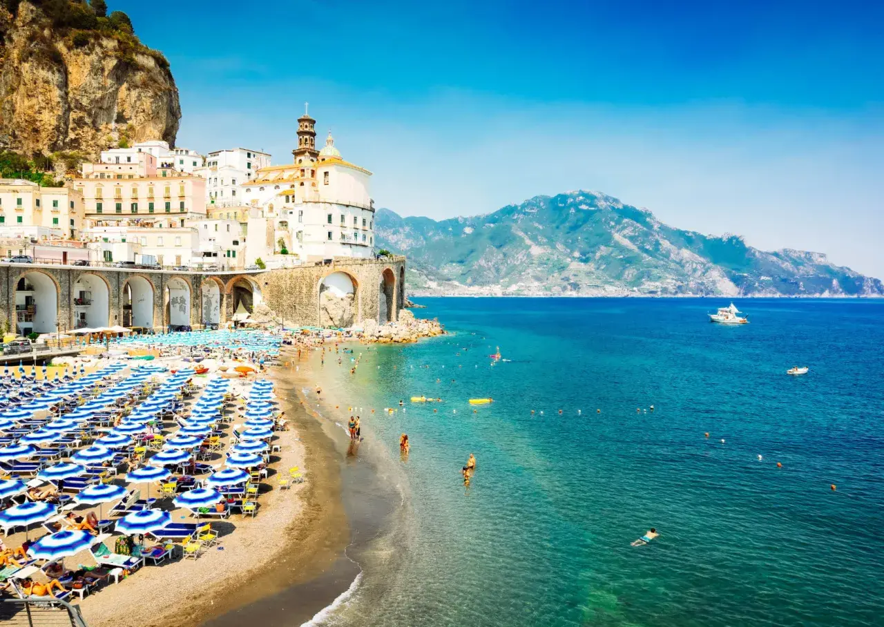 A vibrant beach scene in Naples, with rows of blue umbrellas and sunbathers. Buildings cling to a cliffside, overlooking the sparkling turquoise sea.