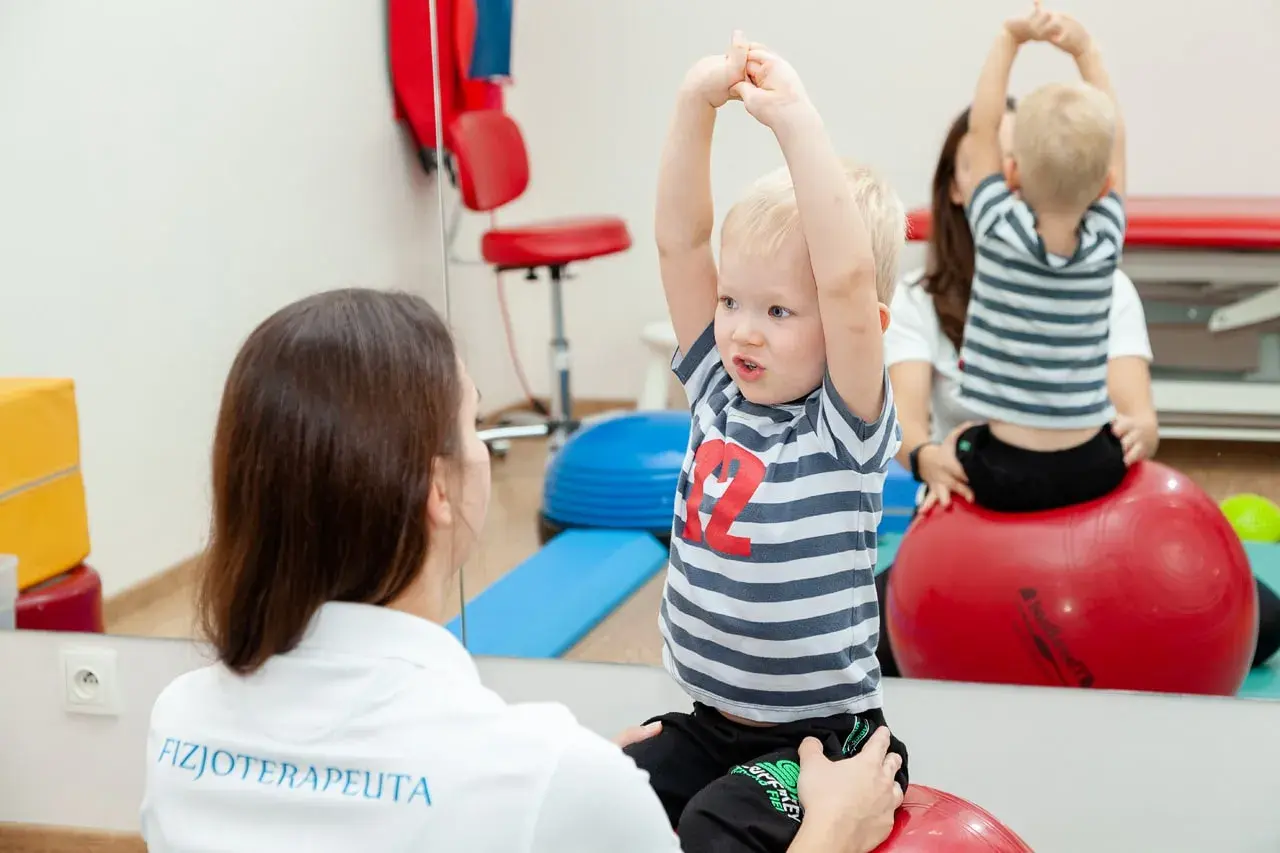 Fizjoterapeutka wspiera chłopca z zaburzeniami rozwojowymi podczas ćwiczeń na piłce rehabilitacyjnej.