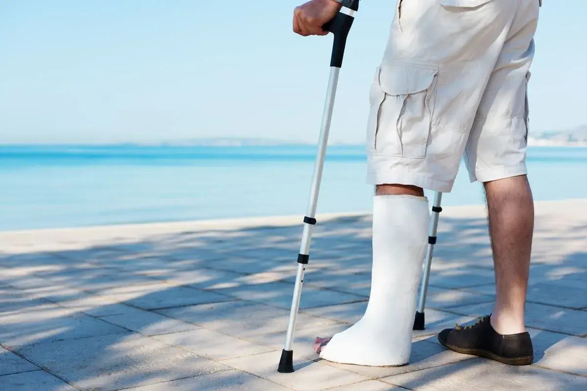 Mężczyzna z białym opatrunkiem na nodze, zamiast gipsu przy złamaniu, wspiera się na kuli, spacerując brzegiem morza.