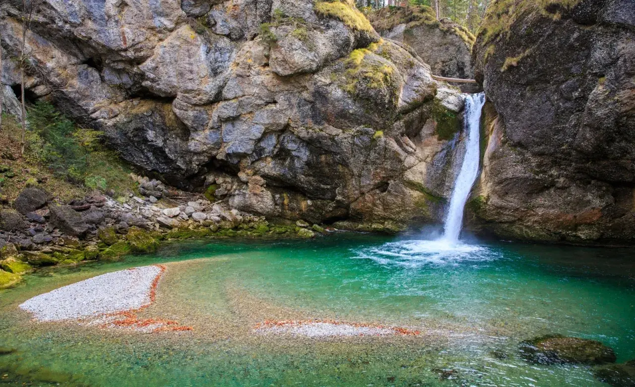 Türkiser bergsee mit einem tosenden wasserfall, der in die klare Tiefe stürzt. Felsen und Moos umrahmen dieses Naturschauspiel.